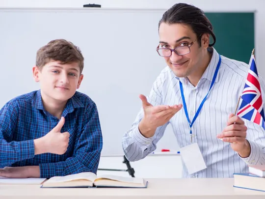Smiling English teacher holding a UK flag next to a student giving a thumbs up in a classroom.