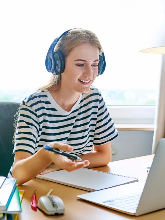 a young female student wearing headphones and a striped shirt, smiling while studying at a laptop for an online class at a desk.