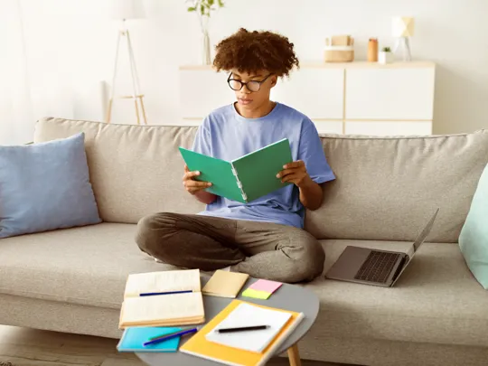 Teenager studying at home, surrounded by textbooks and a laptop, demonstrating the independent learning environment fostered by homeschooling for secondary students.