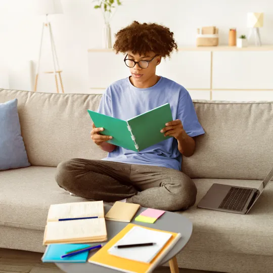 Teenager studying at home, surrounded by textbooks and a laptop, demonstrating the independent learning environment fostered by homeschooling for secondary students.