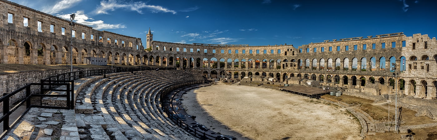 Wide-angle view of a well-preserved Roman amphitheatre with stone seating and arched walls under blue sky.