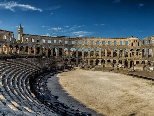 Wide-angle view of a well-preserved Roman amphitheatre with stone seating and arched walls under blue sky.