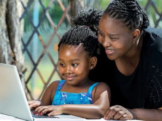 dult assisting a young girl with her studies on a laptop outdoors, as part of their South Africa home education programme.