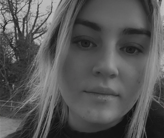 A close up, black and white portrait of a young woman looking directly at the camera, with blurred trees in the background.